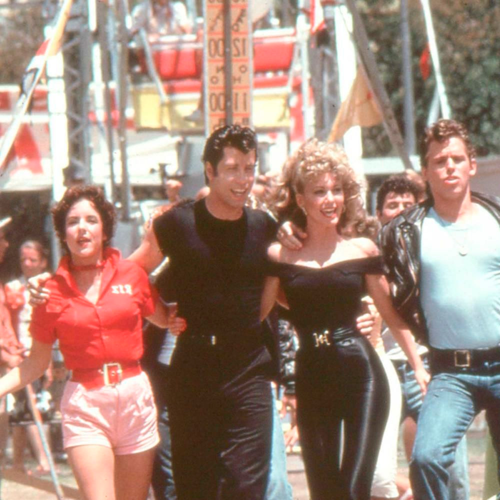 Four people dressed in 1950s-style outfits walk arm-in-arm, smiling, at a carnival or fairground with rides and colorful decorations in the background.