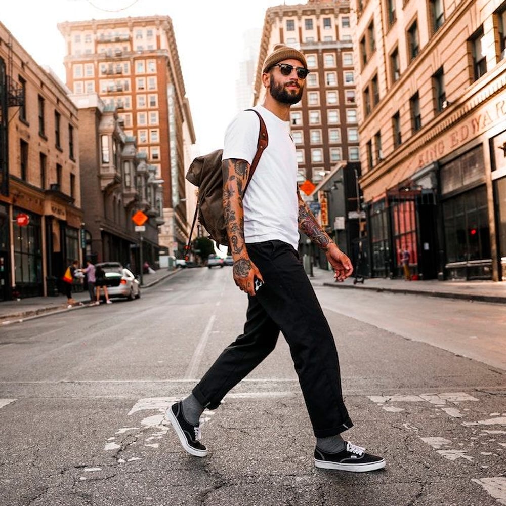 A man with tattoos, sunglasses, and a beanie walks confidently across a city street. He wears a white t-shirt, dark pants, black sneakers, and carries a brown backpack. Tall buildings line the street under a bright sky.