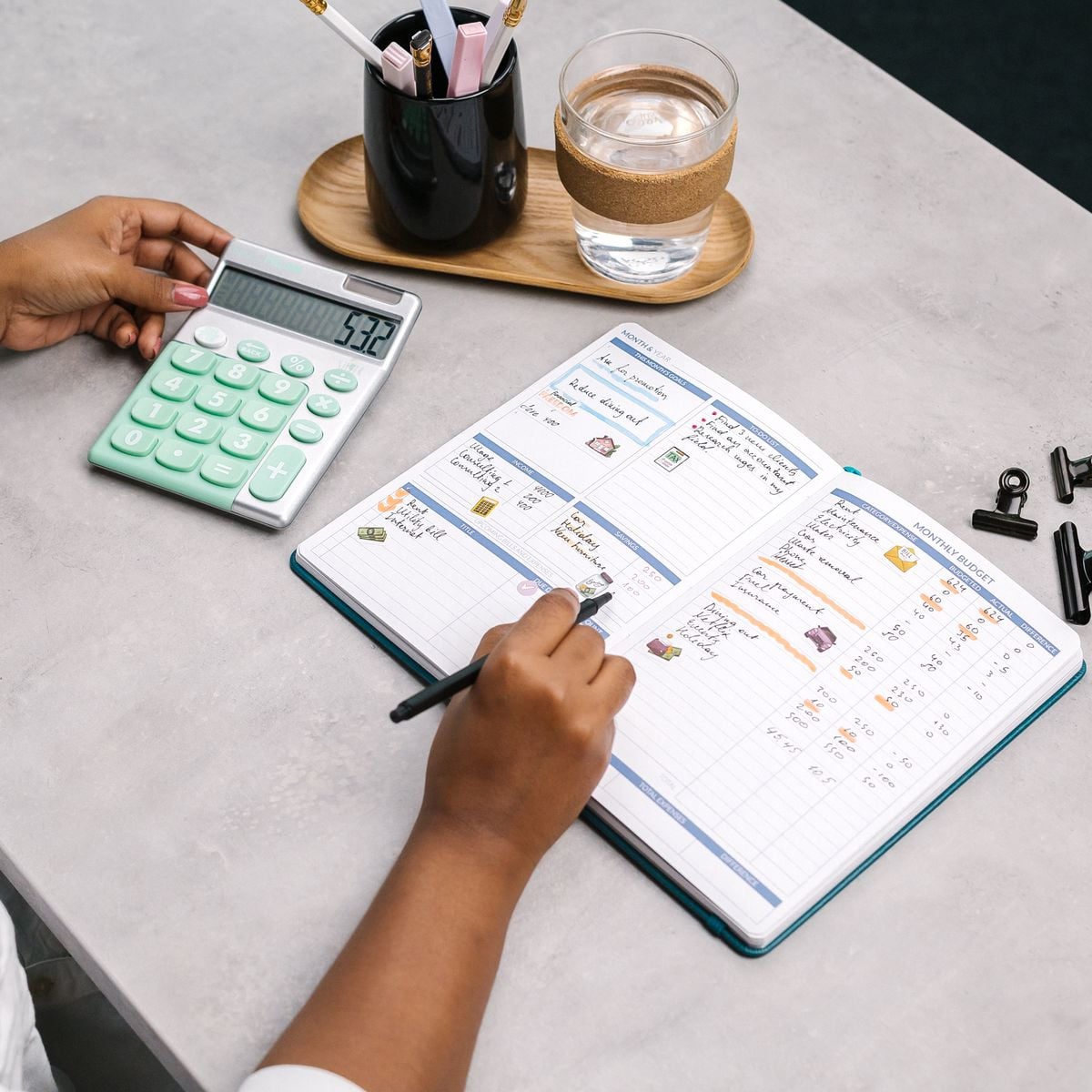 A person writes in a planner with a pen, next to a large green calculator, a glass of water, and a cup holding pens and pencils on a gray desk.