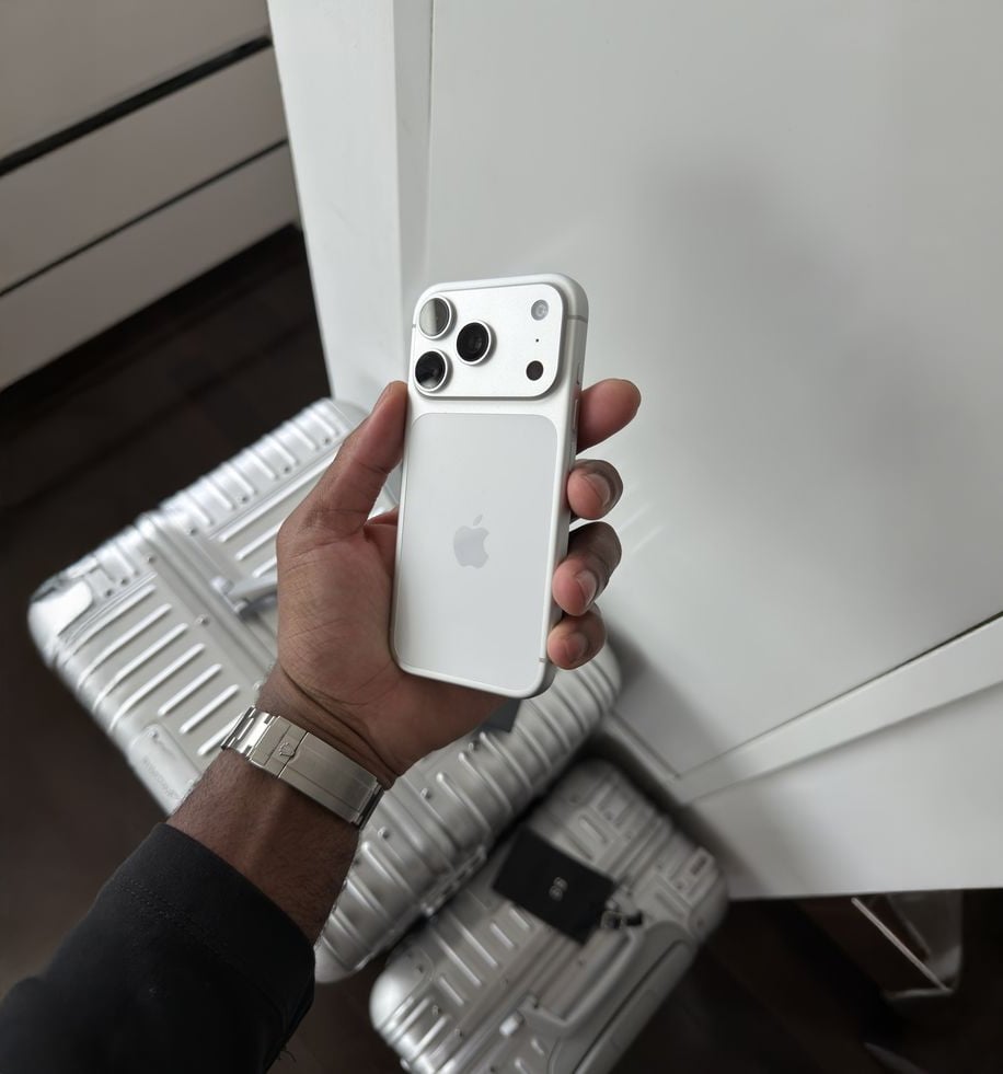 A person wearing a silver watch holds a white iPhone with a case, standing near metallic silver suitcases and a white cabinet.