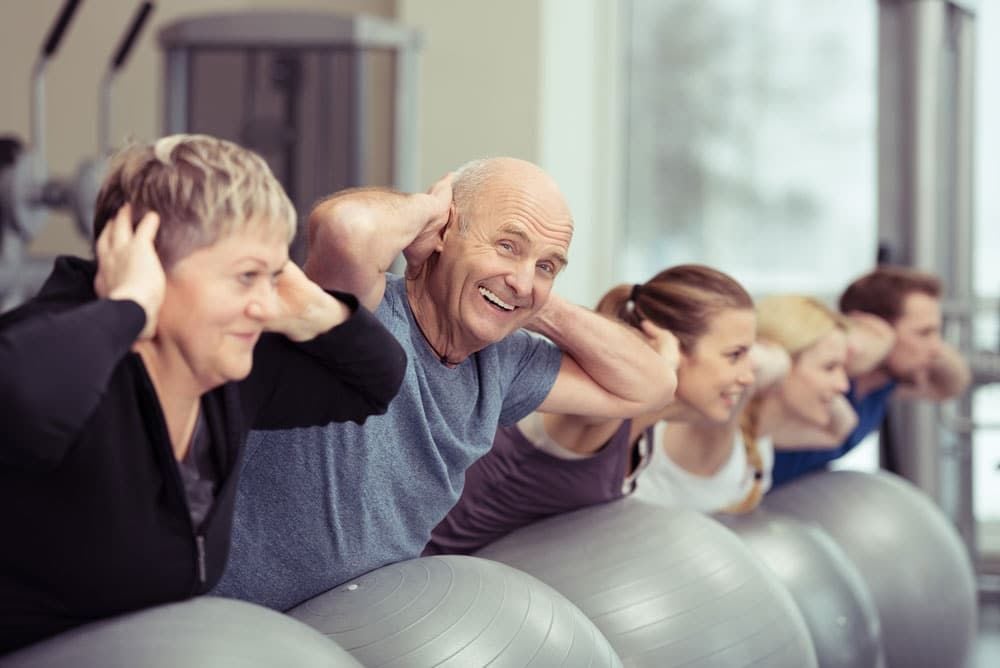 A group of smiling adults, including seniors, do abdominal exercises on fitness balls in a bright gym, each with hands behind their head, lined up side by side.
