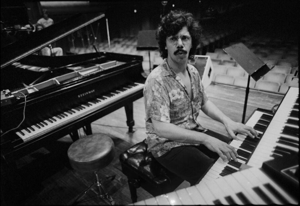 A man with curly hair and a mustache plays a keyboard on stage beside a grand piano. He is seated on a bench, wearing a patterned shirt, and looking toward the camera in an empty auditorium.