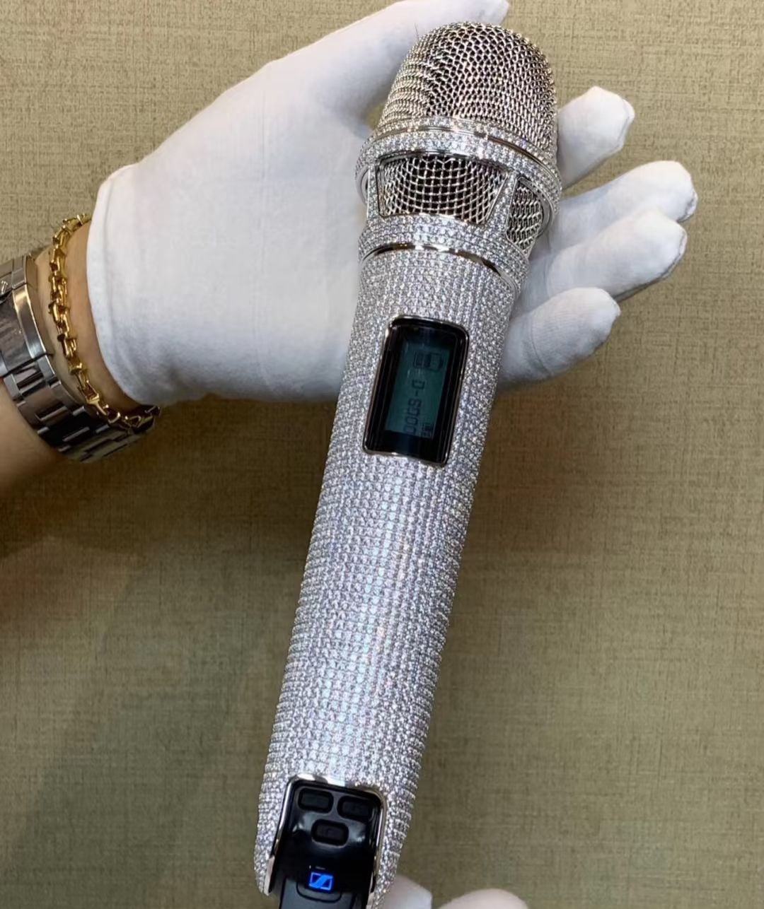 A person wearing white gloves and a gold bracelet holds a wireless microphone encrusted with sparkling crystals against a beige textured background.