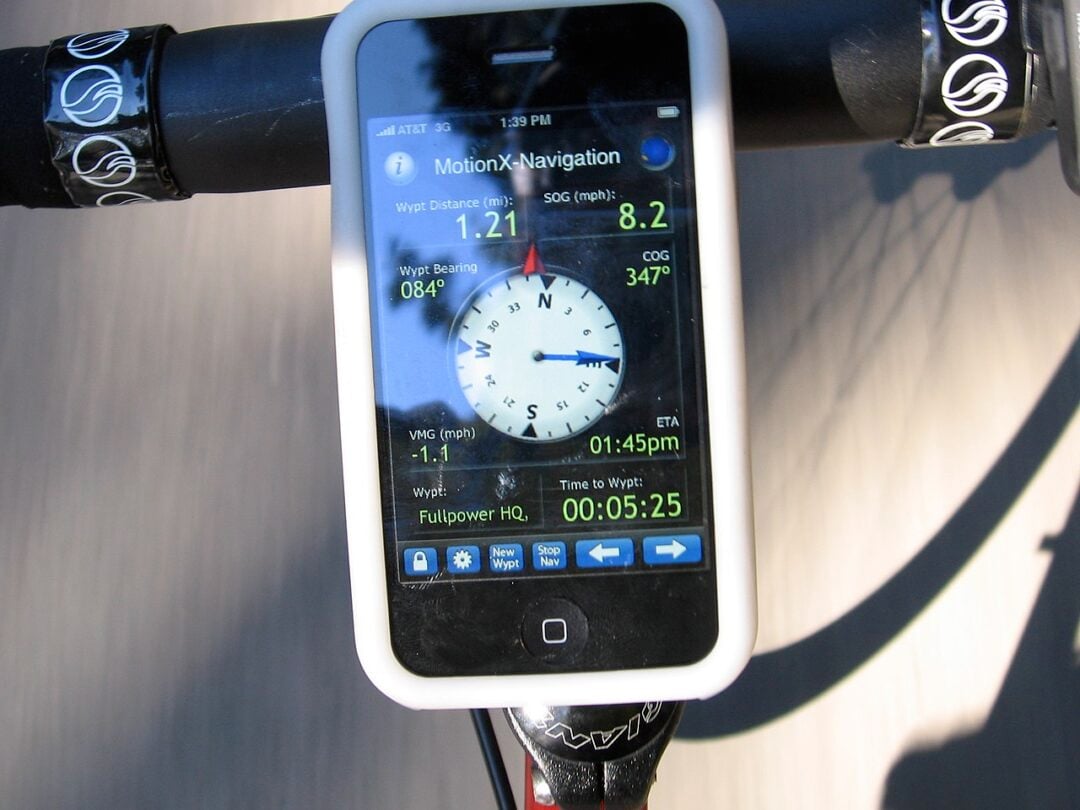 A smartphone mounted on a bicycle handlebar displays a navigation app with a compass, speed, distance, and time information while the bike is in motion.