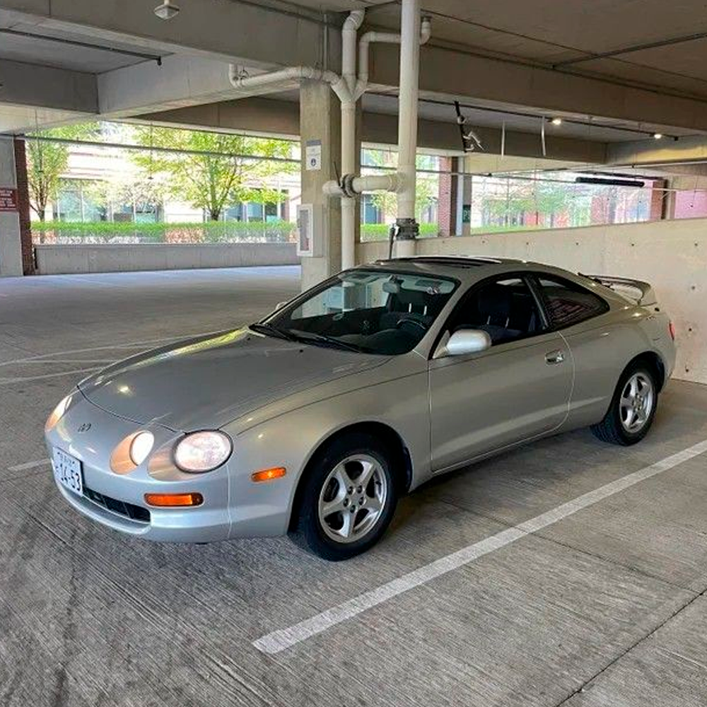 A silver two-door coupe car is parked in a multi-level parking garage, illuminated by natural light from outside. The car's headlights are on, and trees can be seen through the windows in the background.