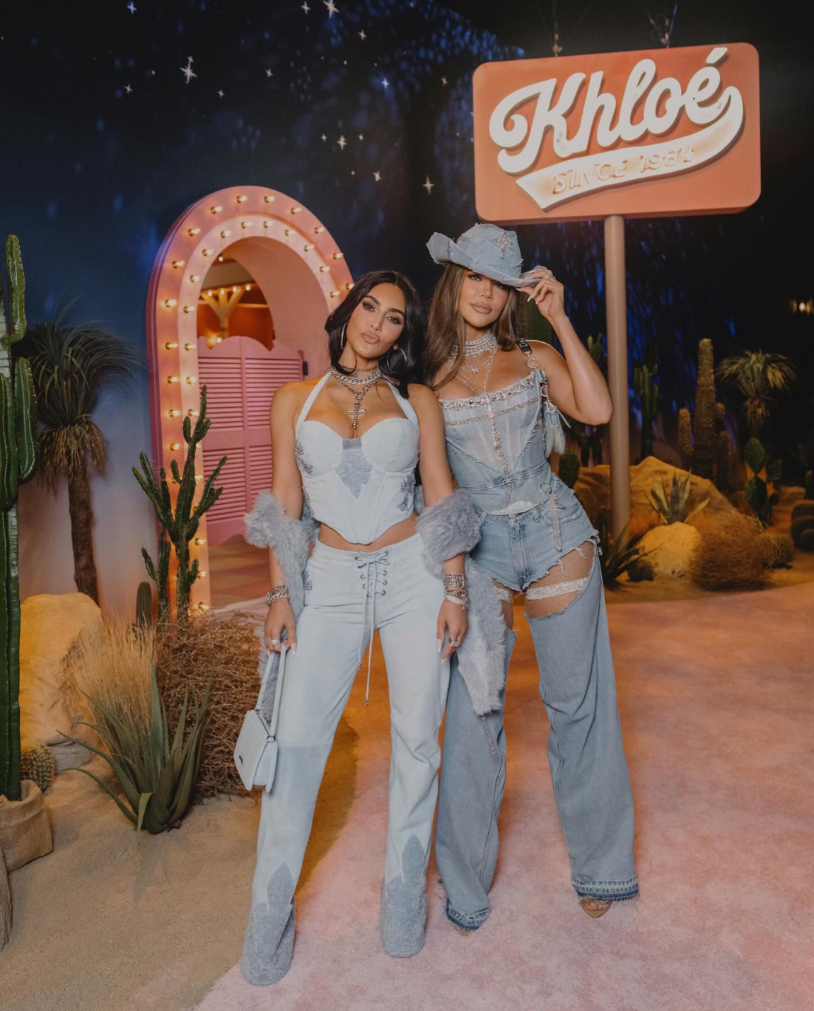 Two women pose together in stylish light denim outfits and fur accents at a western-themed event. They stand on a pink carpet with cacti and a neon sign that reads “Khloé” in the background.