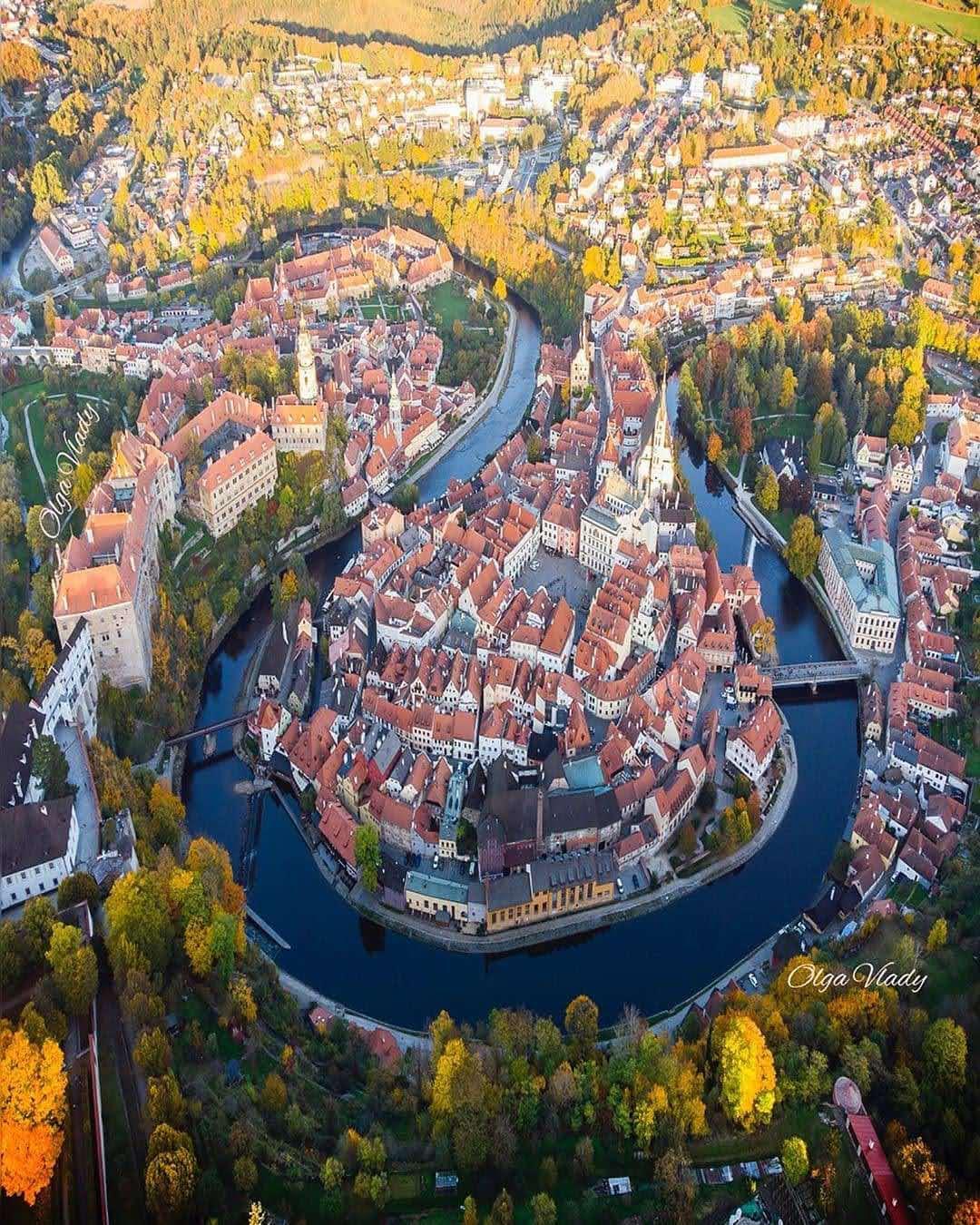 Aerial view of a historic European town with red-roofed buildings surrounded by a winding river and lush greenery, bathed in warm sunlight.