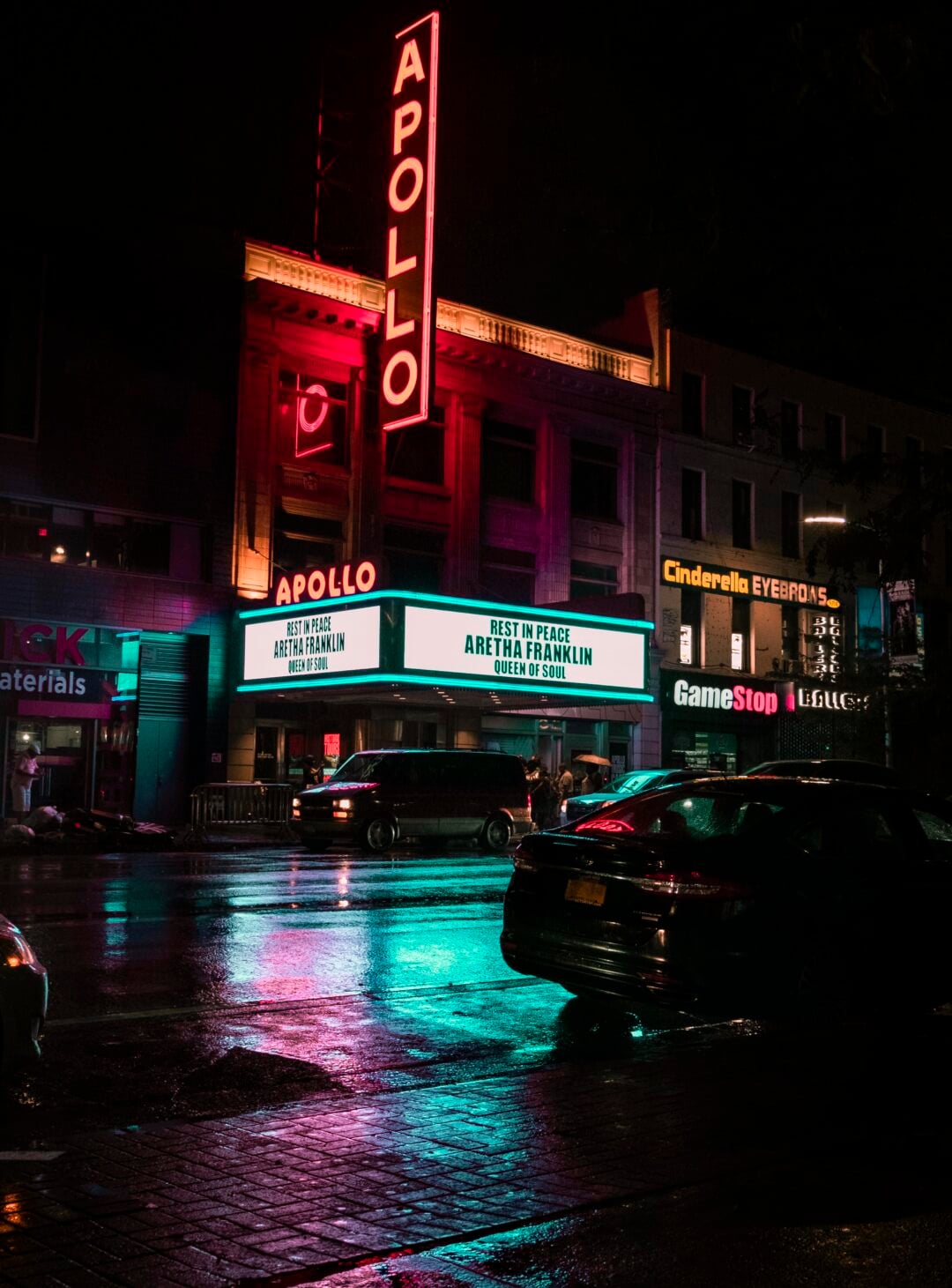The Apollo Theater at night, illuminated by neon lights reflecting on wet pavement. The marquee reads, "Rest in Peace Aretha Franklin Queen of Soul." Cars are parked along the street, and nearby storefronts are visible.