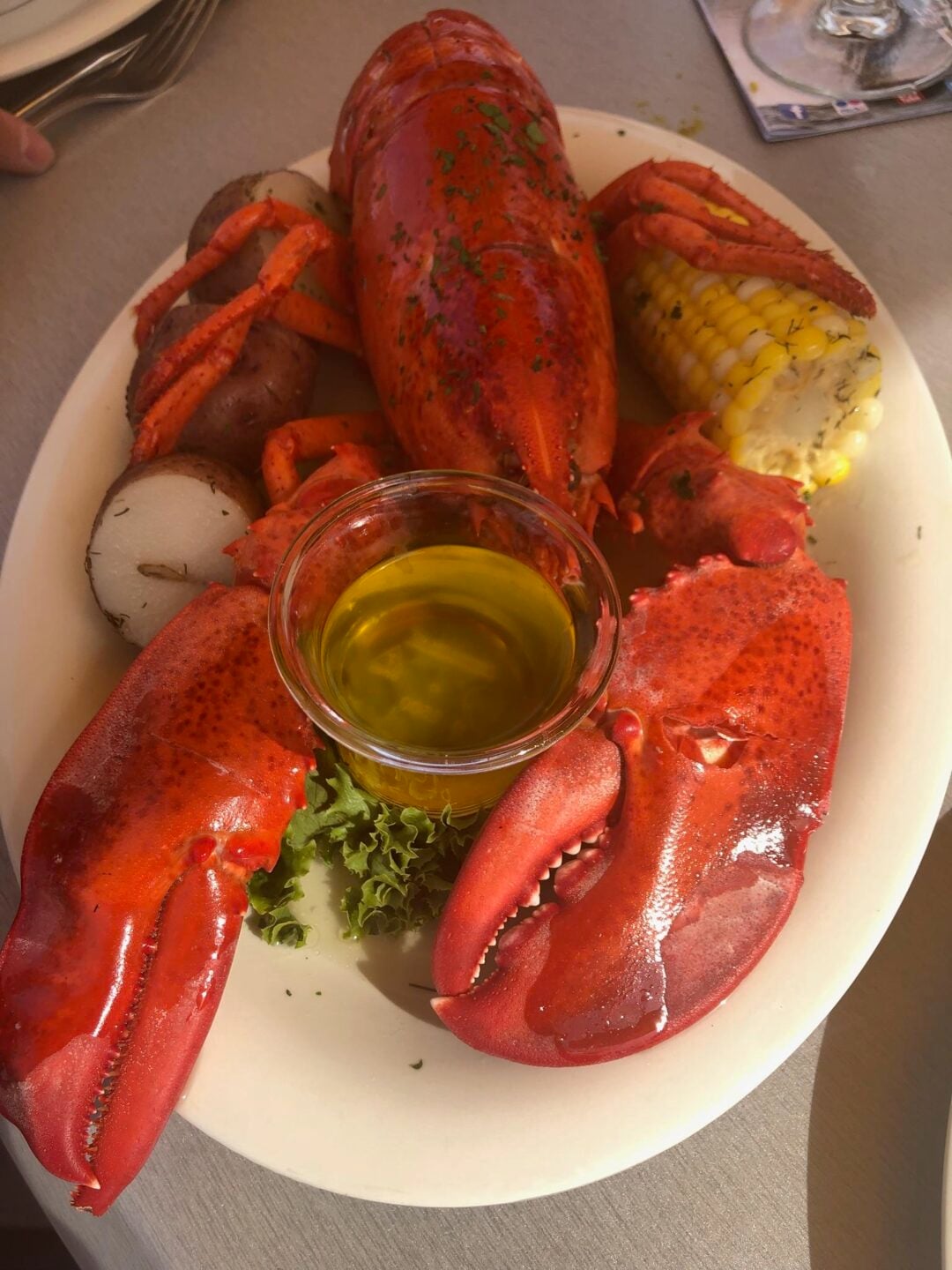 A plate with a cooked lobster, corn on the cob, red potatoes, leafy greens, and a small cup of melted butter.
