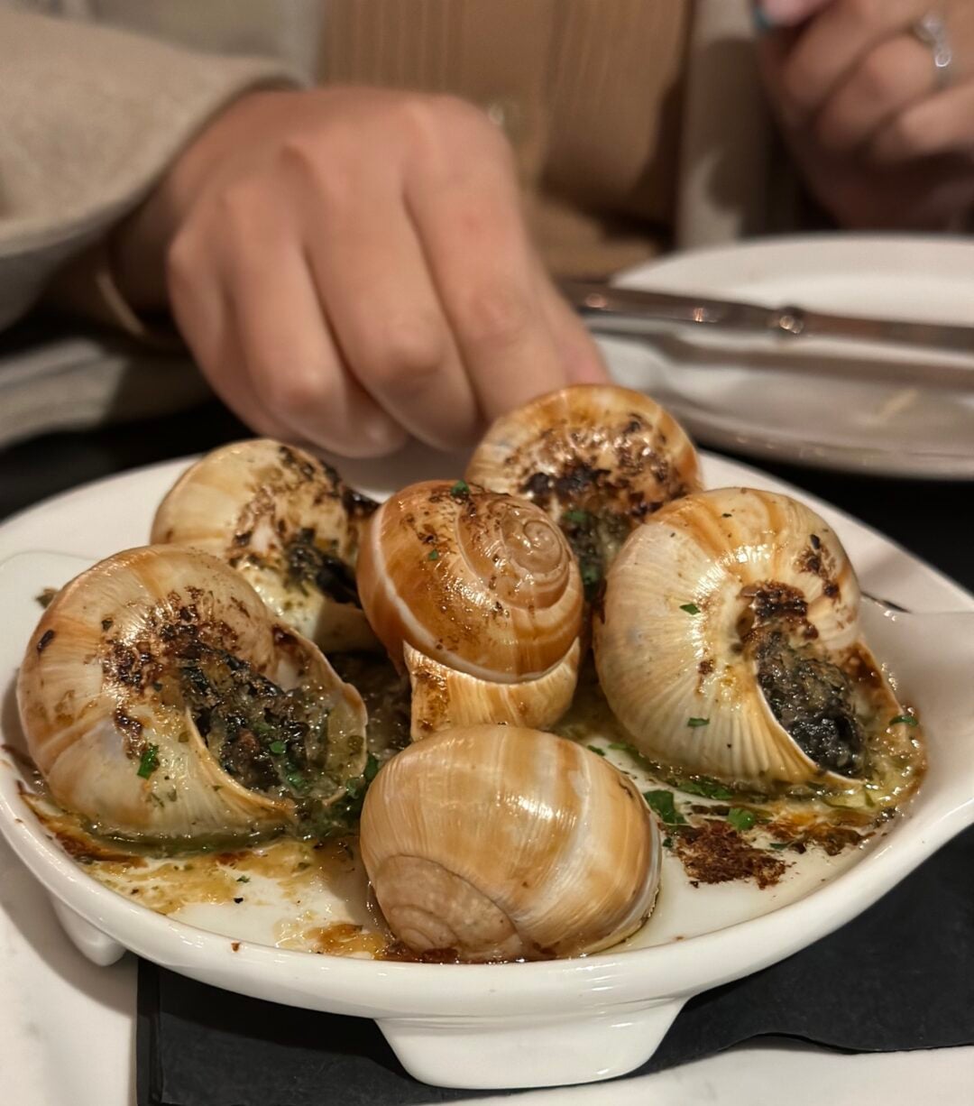 A hand reaches toward a white dish filled with six baked escargots in their shells, topped with herbs and garlic butter. The dish sits on a black napkin, with an empty plate and utensils in the background.