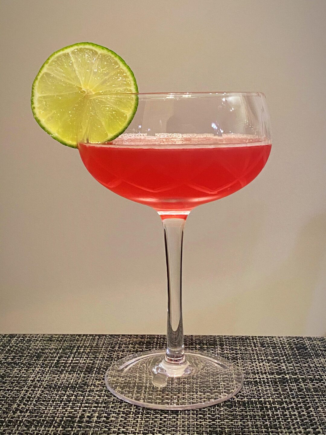 A pink cocktail served in a coupe glass, garnished with a fresh lime wheel on the rim, placed on a textured dark surface against a plain light background.