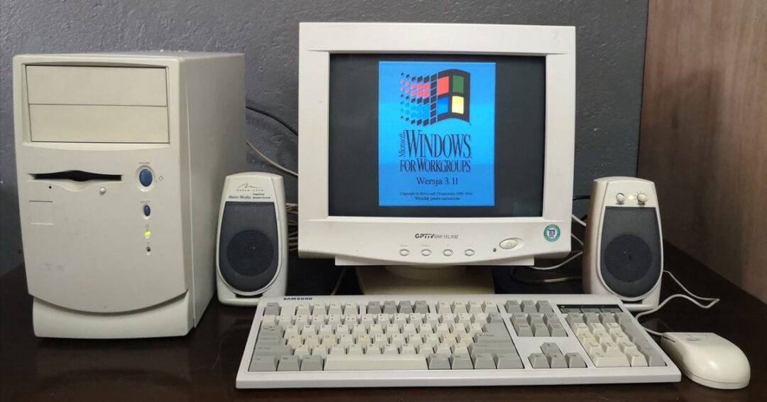 A vintage desktop computer setup with a beige tower, CRT monitor displaying a Windows for Workgroups 3.11 startup screen, two speakers, and a mechanical keyboard on a dark desk.