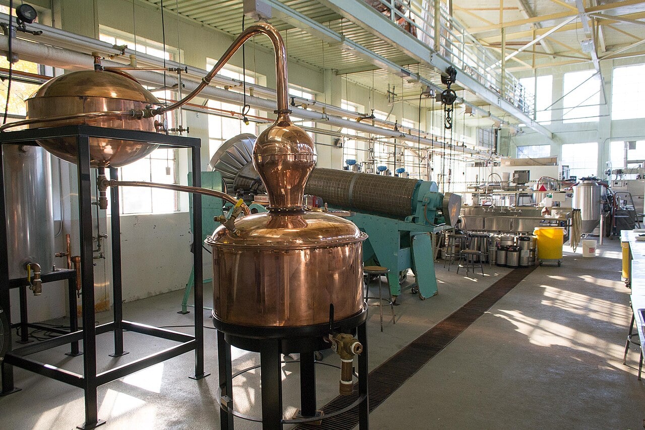 A bright, spacious industrial facility with large distillation equipment, including a shiny copper still in the foreground and various metal machinery and containers arranged throughout the area.