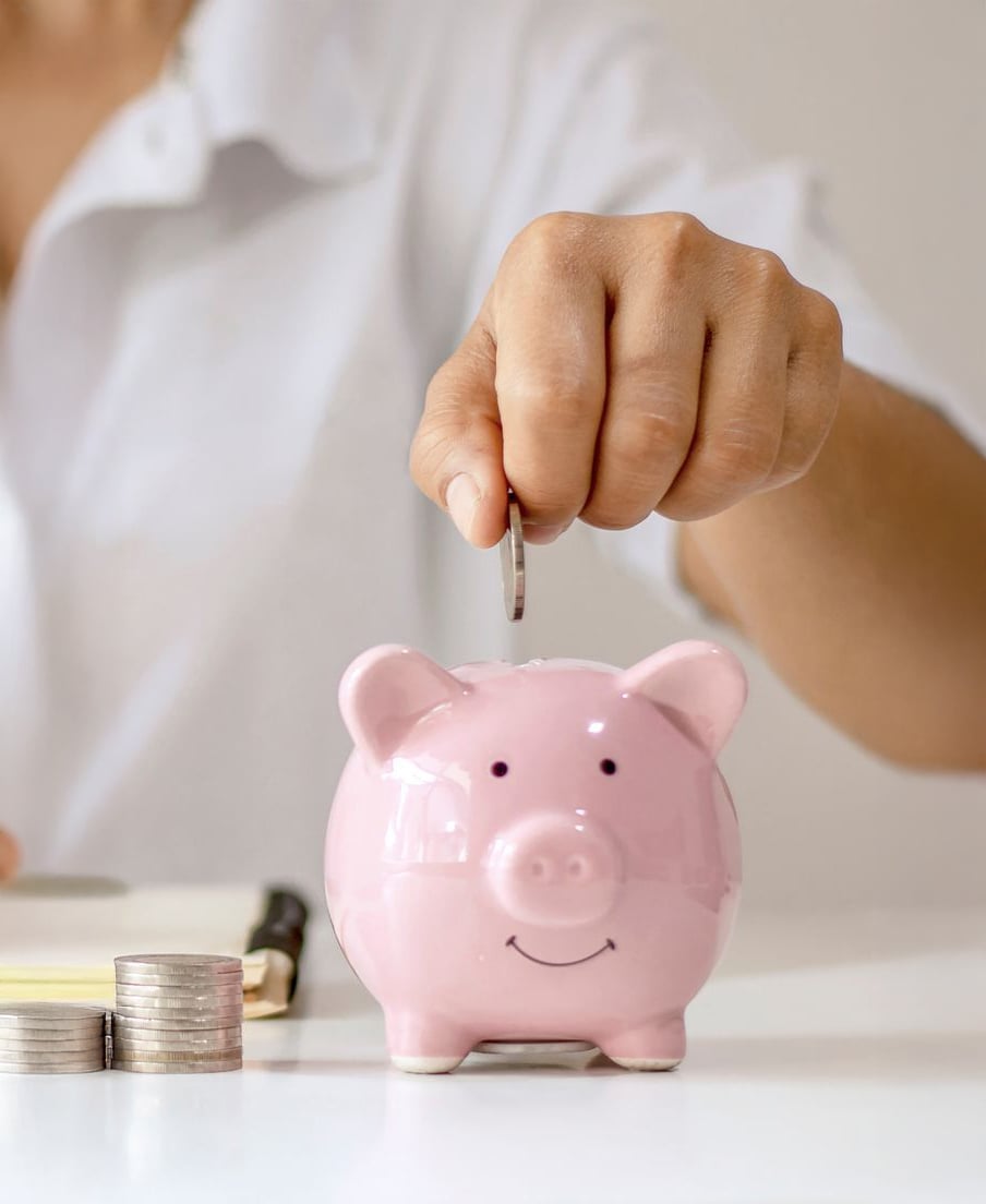 A person drops a coin into a pink piggy bank. Stacks of coins sit nearby, and an open notebook is partially visible in the background. The scene suggests saving money or financial planning.