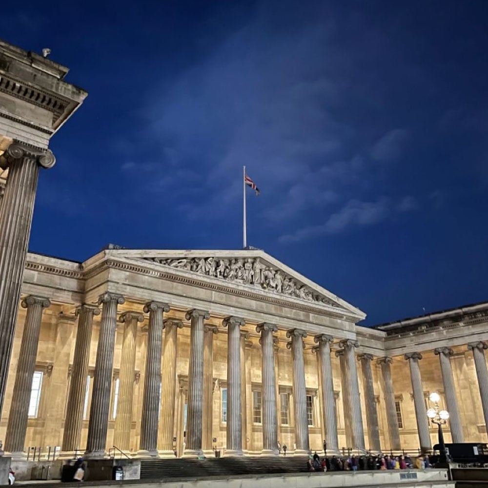 A grand neoclassical building with tall columns and a British flag atop the roof, illuminated at night against a deep blue sky. Several people are visible near the entrance.