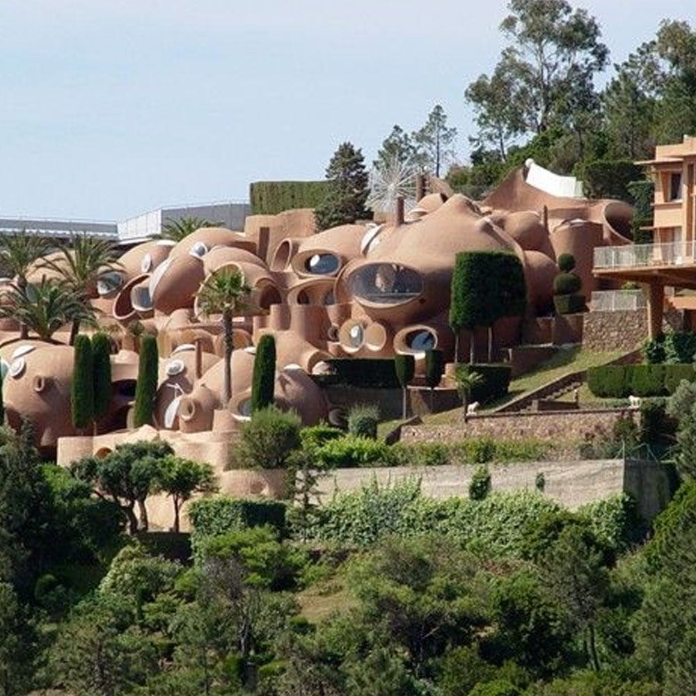 A cluster of unique, rounded, clay-colored buildings with circular windows, surrounded by greenery and trees on a hillside under a clear sky.