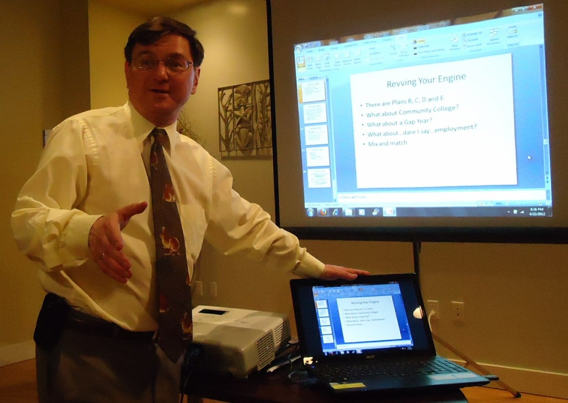 A man in a dress shirt and tie stands beside a projector and laptop, gesturing toward a projection screen displaying a PowerPoint slide titled "Revving Your Engine" in a well-lit room.