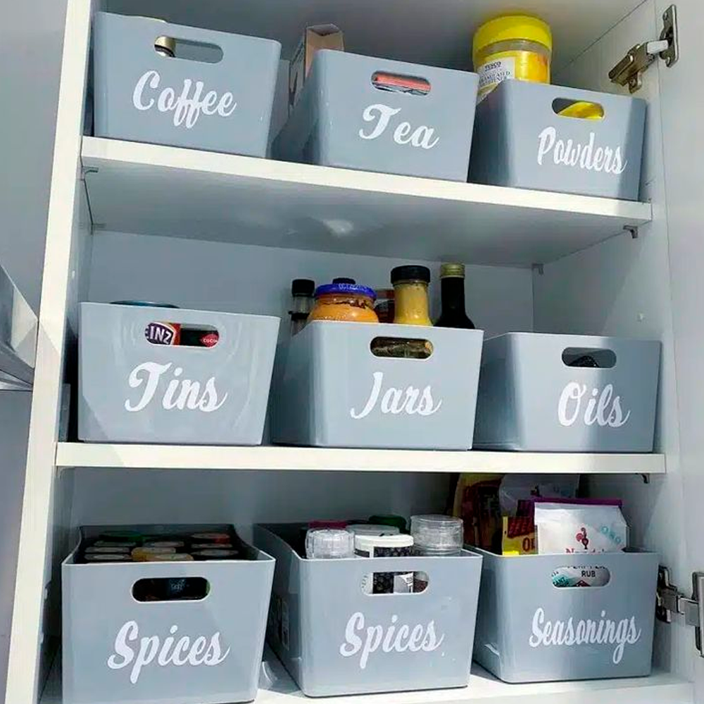Three shelves in a cupboard display labeled grey storage bins for coffee, tea, powders, tins, jars, oils, spices, and seasonings, with various food items organized neatly inside each bin.