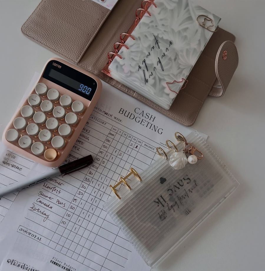 A calculator, pen, planner, and cash budgeting sheet are arranged neatly on a desk, along with an envelope-style pouch labeled "savings," gold paper clips, and a decorative keychain.