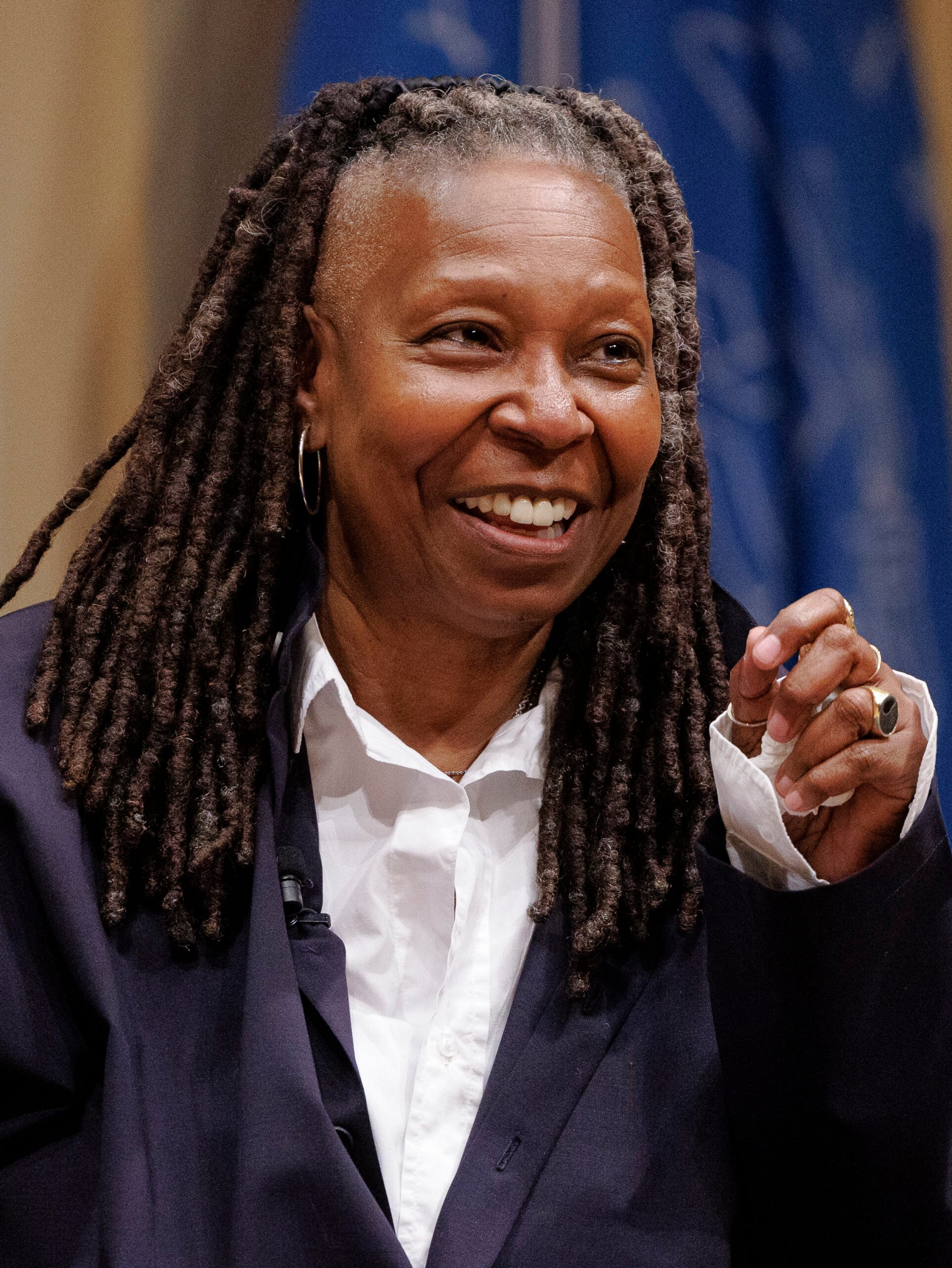 A smiling woman with long, dark dreadlocks wearing a white shirt and dark blazer, gesturing with her hand, stands in front of a blurred blue and beige background.