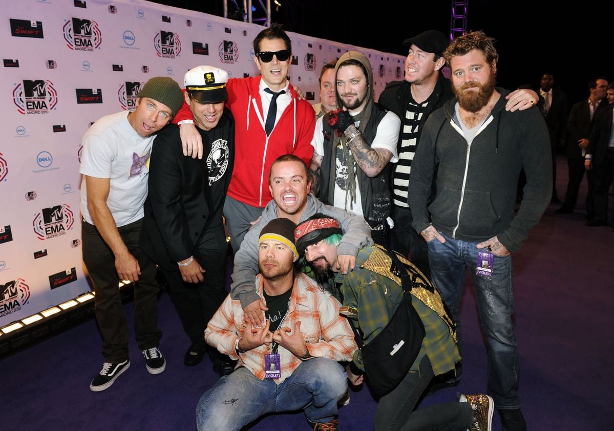 A group of ten men pose together on a purple carpet at an event, standing and kneeling in front of a backdrop covered with MTV and EMA logos. They are smiling and wearing casual or eclectic outfits.