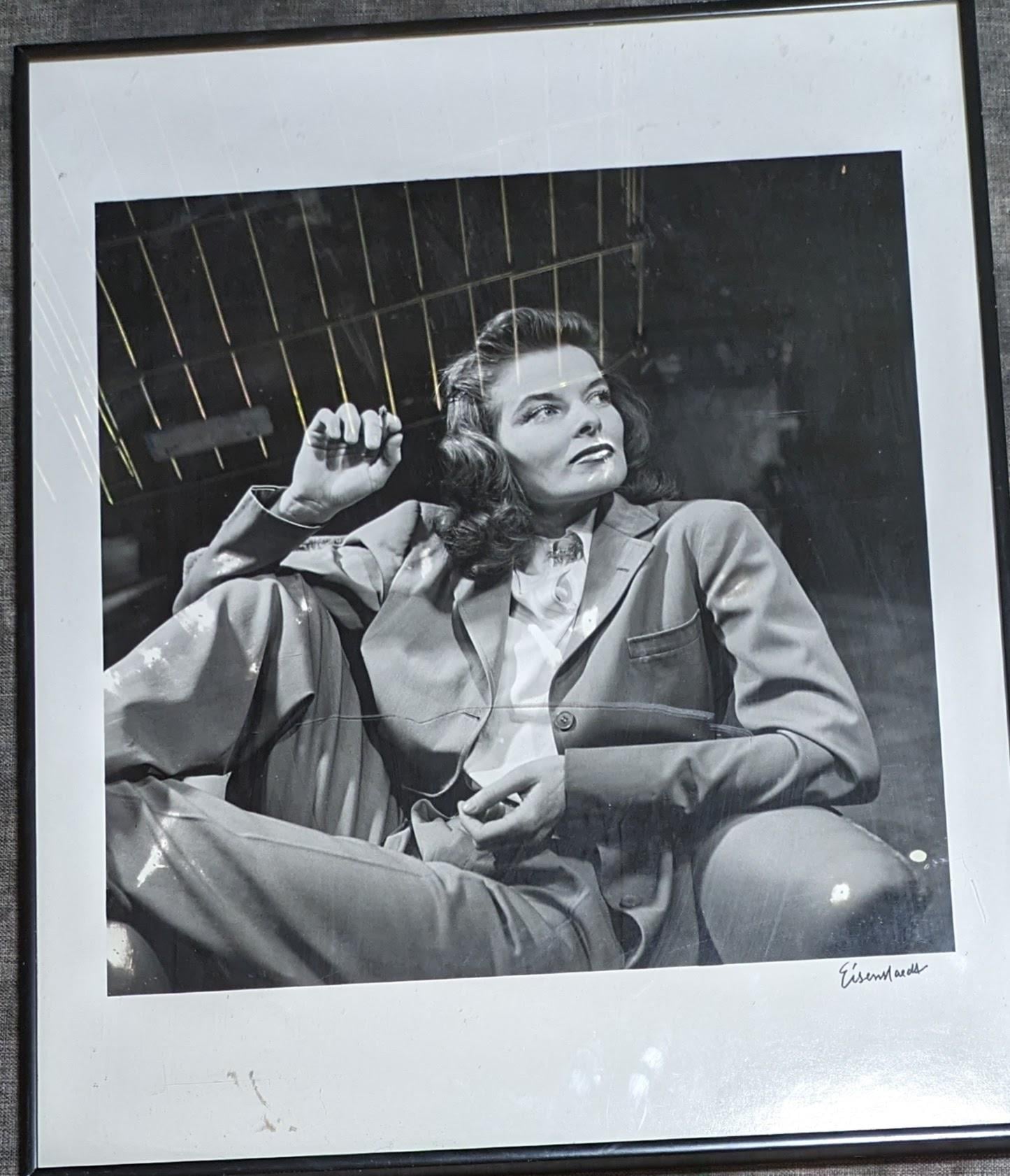 A black-and-white photo of a woman in a suit sitting relaxed in a chair, leaning back with one arm raised. She has wavy hair and a confident expression. The image is in a simple black frame and signed in the corner.