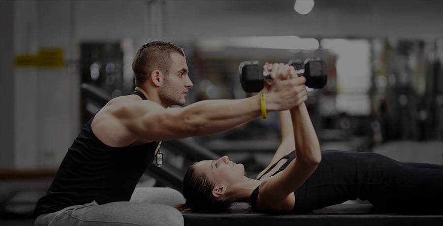 A man assists a woman with a dumbbell chest press exercise as she lies on a bench in a gym. The man is spotting her, offering support and guidance during the workout.