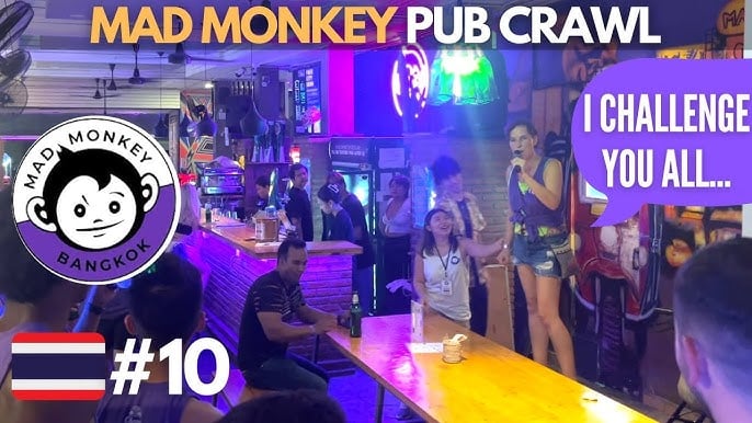 A lively bar scene shows people gathered for the Mad Monkey Pub Crawl in Bangkok. A woman stands on a table with a microphone, addressing the crowd. The Thai flag and the number 10 are visible in the foreground.