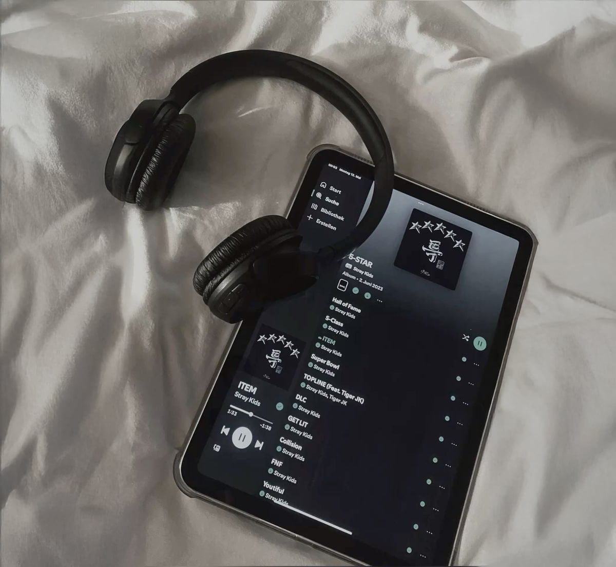 A pair of black headphones rests on a tablet displaying a music playlist on a streaming app, all placed on a white, wrinkled bedsheet.
