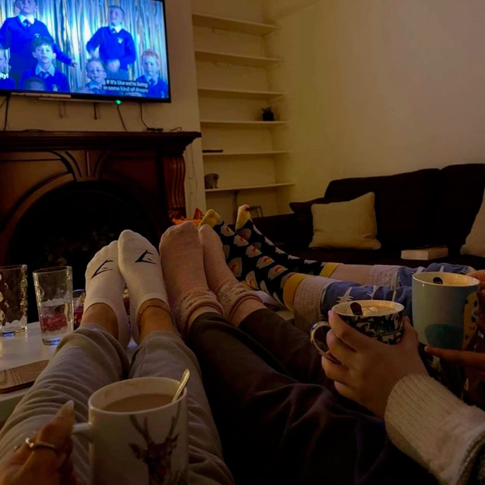 Four people relax on a couch with their feet up on a table, wearing cozy socks and holding mugs. An empty shelf, cushions, and a TV showing a show are visible in the warmly lit living room.