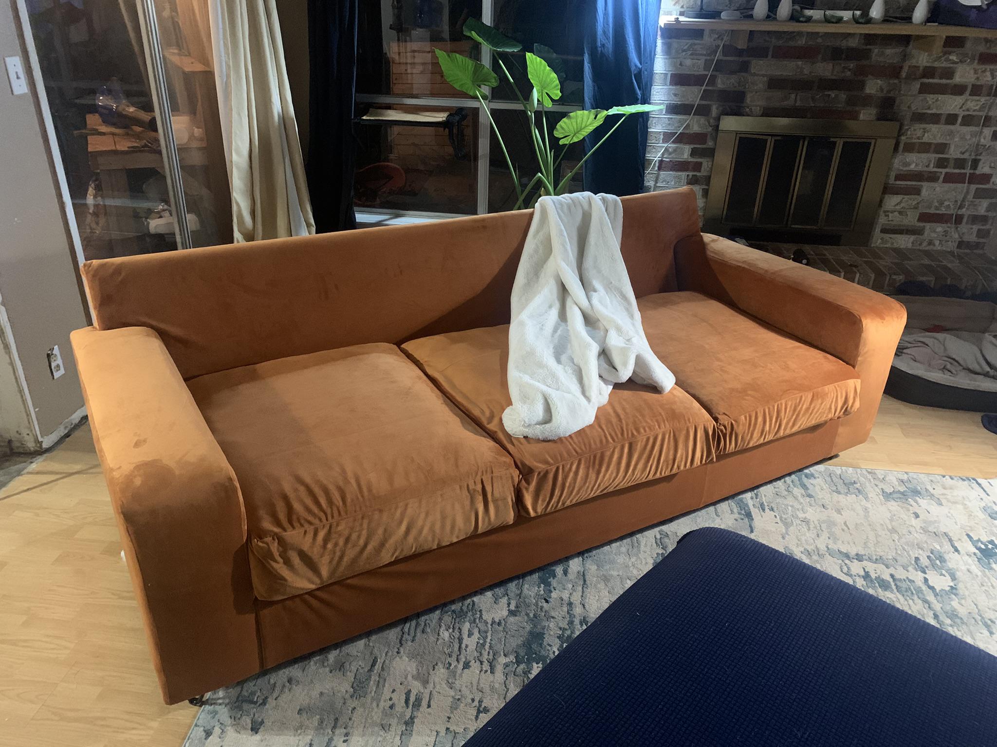 A rust-colored velvet sofa with three seat cushions is draped with a white blanket. The sofa sits on a blue and white rug in a cozy living room with a fireplace and potted plant in the background.