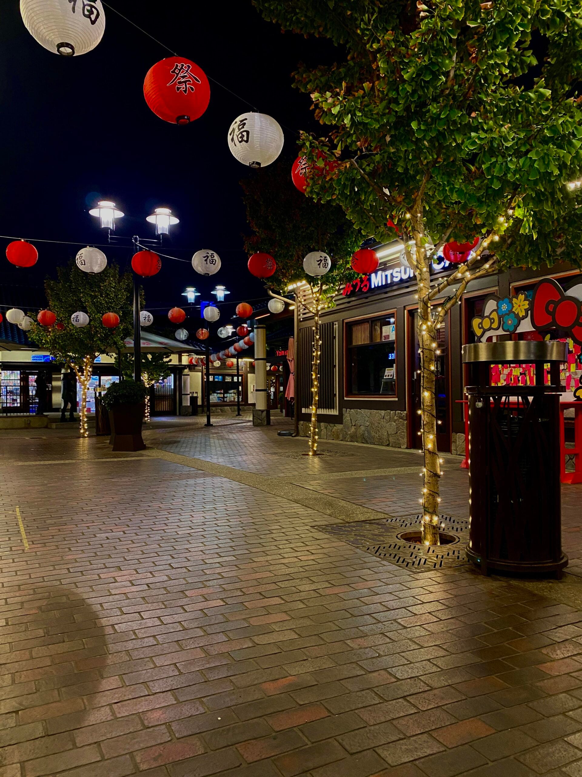 A brick-paved outdoor walkway at night is decorated with red and white lanterns hanging above. Trees wrapped in string lights line the path near shops, including a Japanese restaurant and a Hello Kitty display.