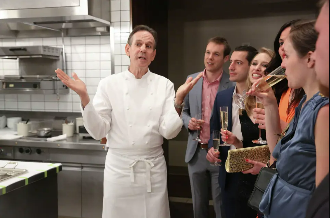A chef in a white uniform gestures while speaking to a group of well-dressed people holding champagne glasses in a modern kitchen setting.