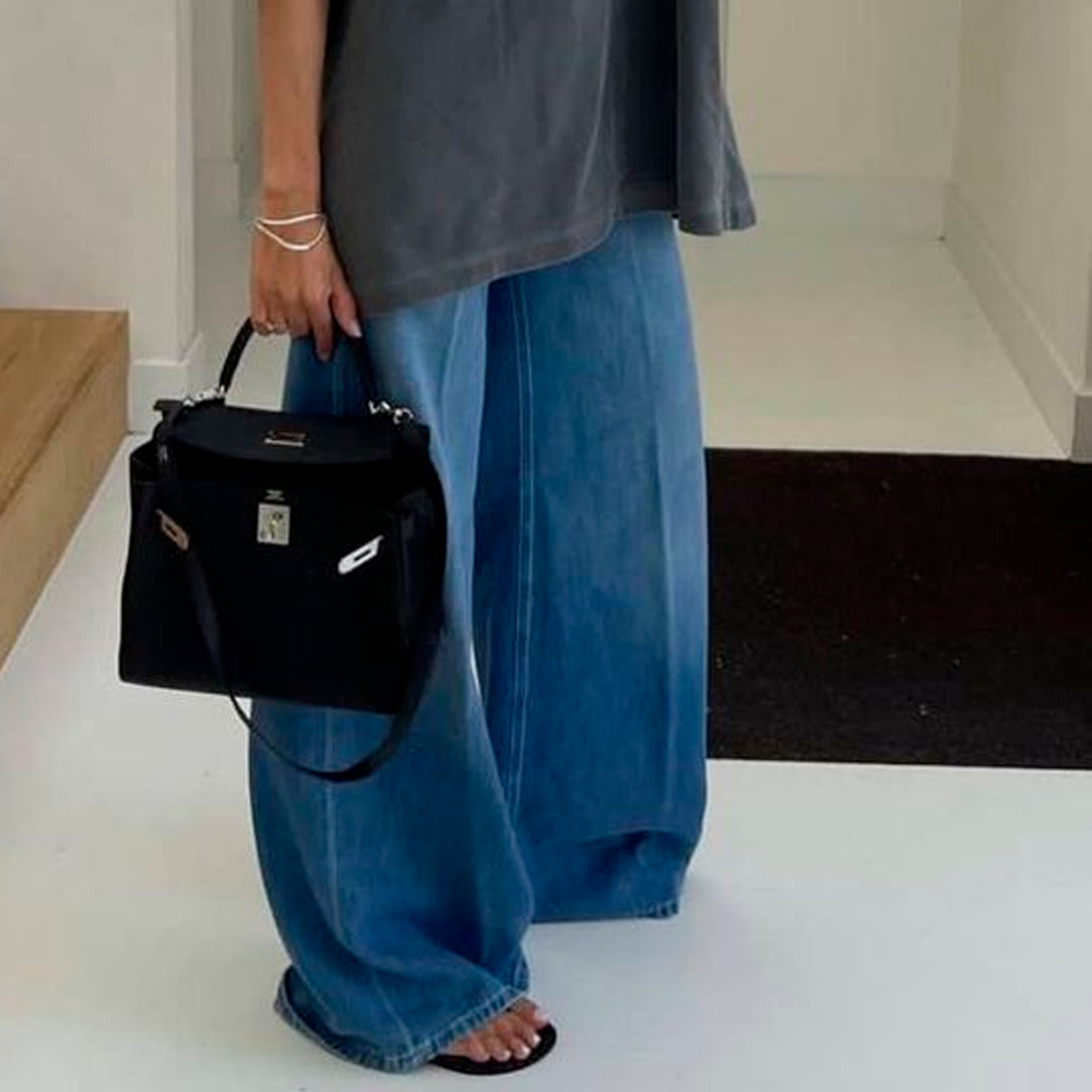 A person wearing wide-leg blue jeans, a loose grey top, black sandals, and holding a black handbag stands indoors on a white floor next to wooden stairs.