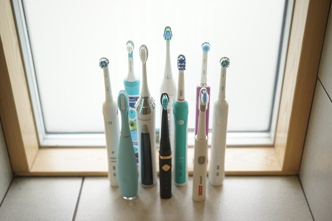 A group of ten electric toothbrushes in various colors and designs are standing upright on a tiled floor in front of a frosted window with a wooden frame.