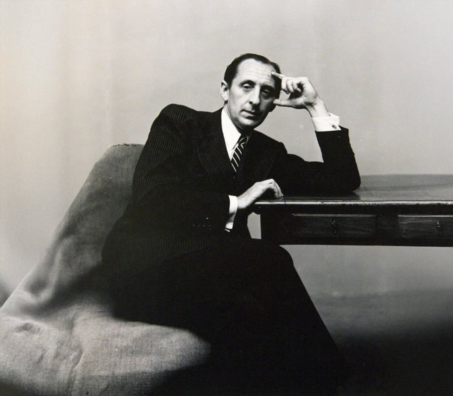A man in a pinstripe suit sits on a draped chair, leaning his head on his hand with his elbow resting on a wooden table, gazing thoughtfully at the camera against a plain background.