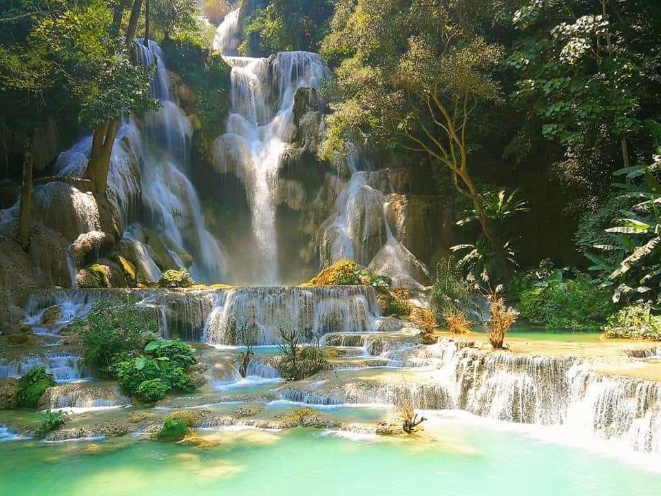 A multi-tiered waterfall cascades over rocks into turquoise pools, surrounded by lush green trees and foliage in bright sunlight.