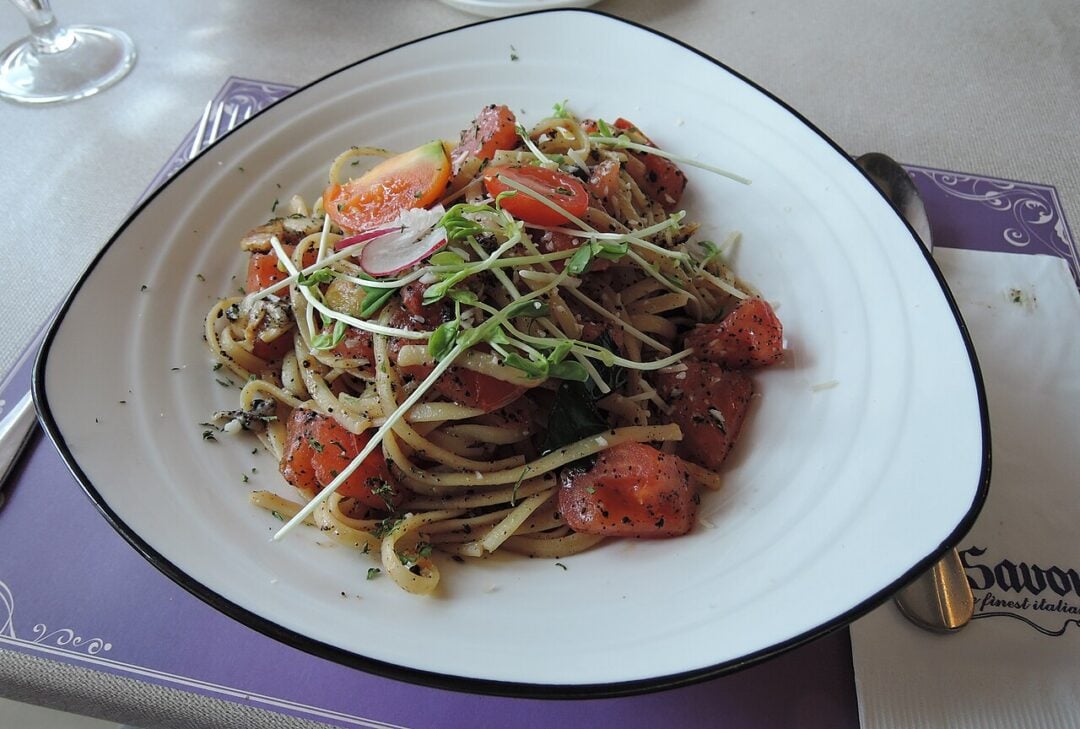 A plate of pasta topped with chopped tomatoes, fresh greens, and herbs is served on a white dish, set on a placemat with a napkin, cutlery, and a bread basket nearby on a table.
