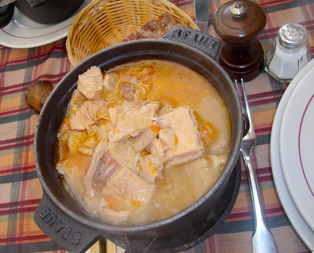 A black Staub pot filled with a stew containing chunks of meat, set on a plaid tablecloth next to a pepper grinder, salt shaker, bread basket, and a plate with cutlery.