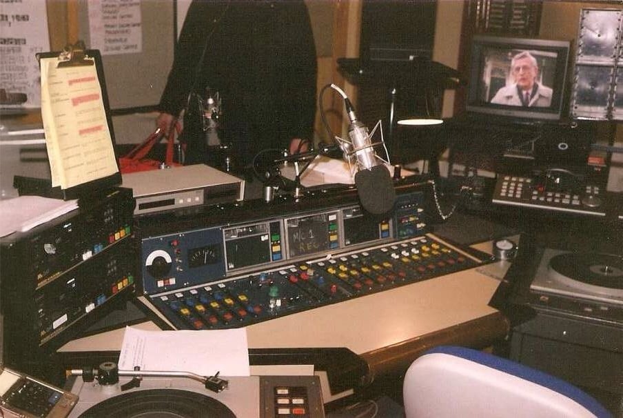 A radio broadcast studio with a large mixing console, microphones, various audio equipment, and a TV displaying a person wearing a suit and hat. Papers and headphones are scattered on the desk.