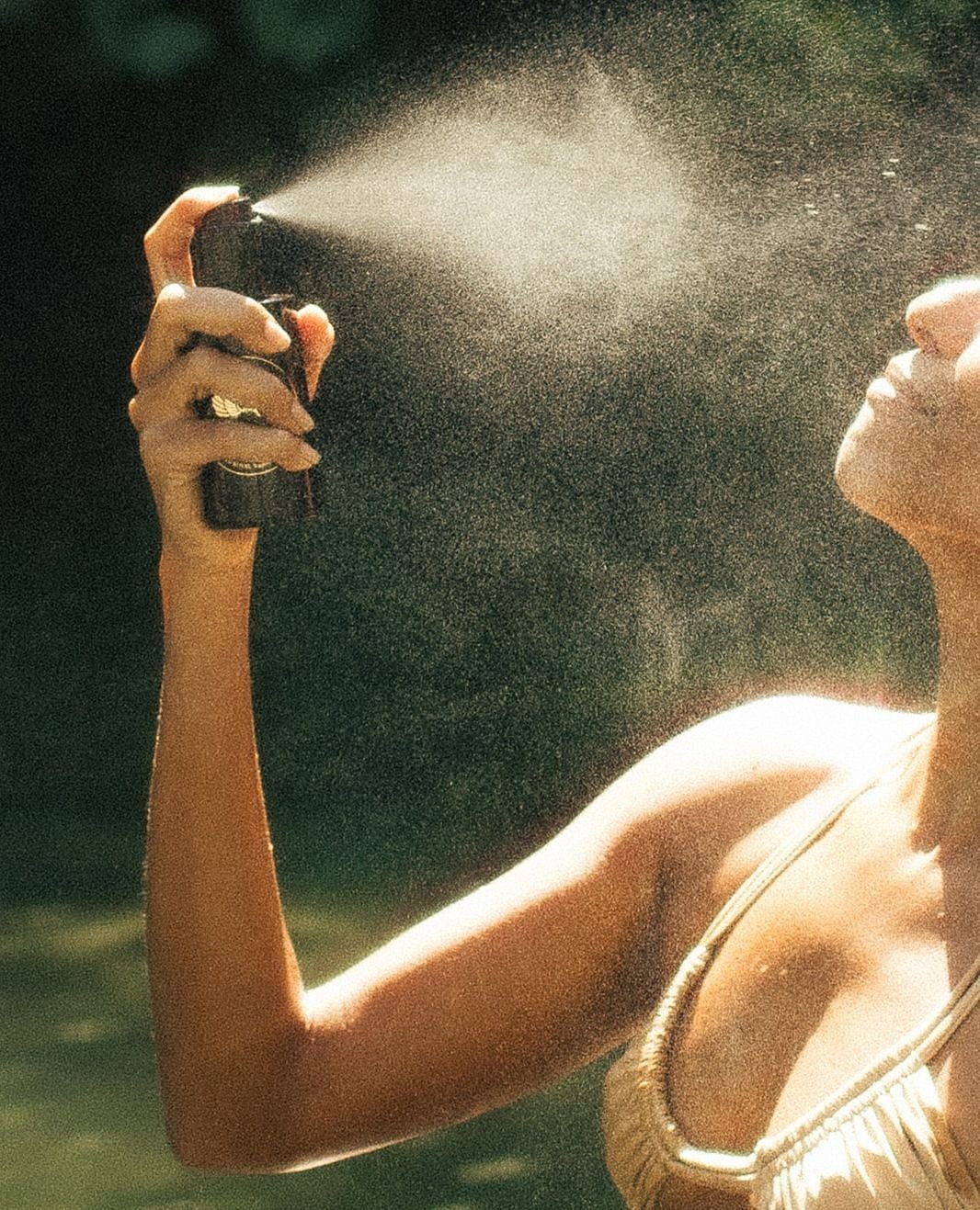 A person in a sleeveless top sprays mist from a bottle onto their face outdoors, with sunlight highlighting the fine spray and their skin.