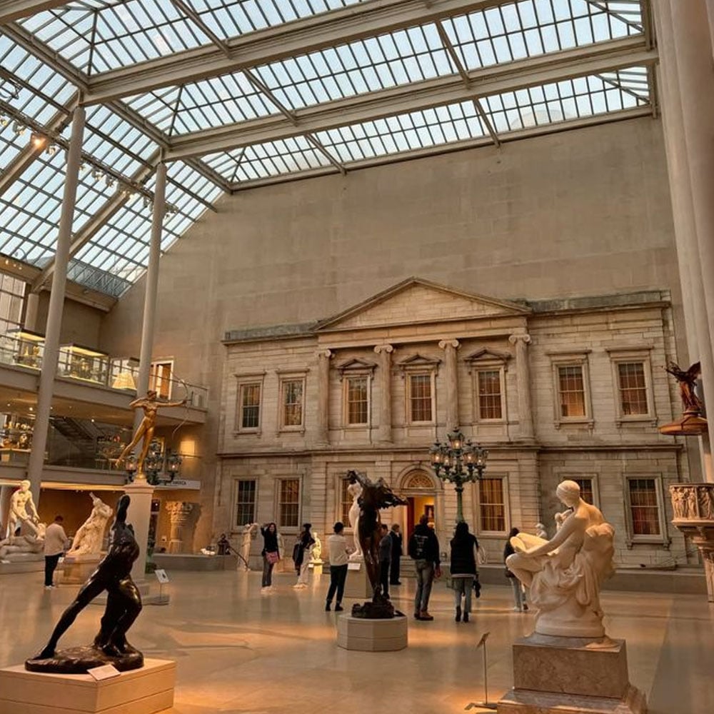 A spacious museum hall with a glass ceiling is filled with classical statues and visitors. In the background stands a grand, columned stone facade reminiscent of historic architecture. Warm sunlight fills the room.