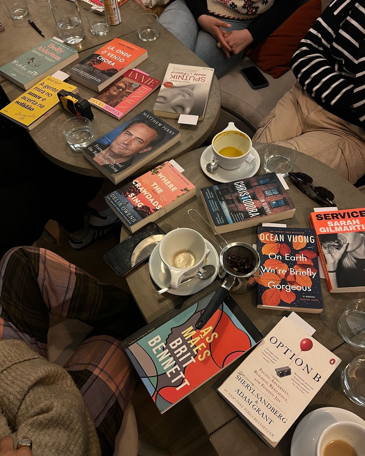 A group of people sit around a table covered with books, drinks, and snacks. The table features diverse books such as "Option B," "As Meat Loves Salt," and "On Earth We’re Briefly Gorgeous.