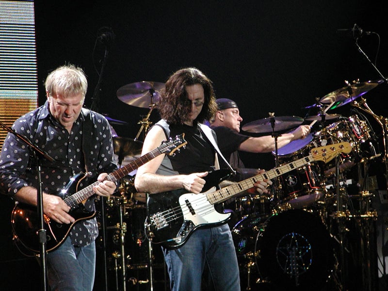 Three musicians perform on stage: one playing electric guitar, one playing bass guitar, and one on drums in the background. All are focused on their instruments under stage lighting.