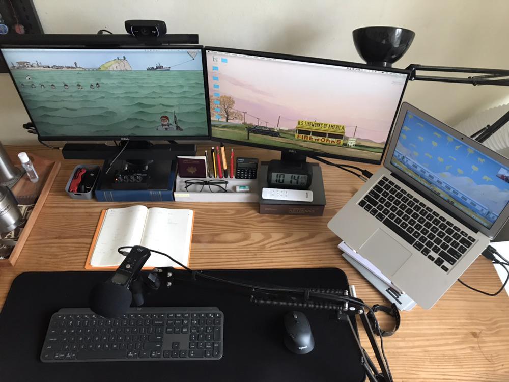 A tidy workspace with a wooden desk features two large monitors, a laptop on a stand, a keyboard, mouse, microphone, notebook, stationery, and speakers, creating an organized and modern office setup.