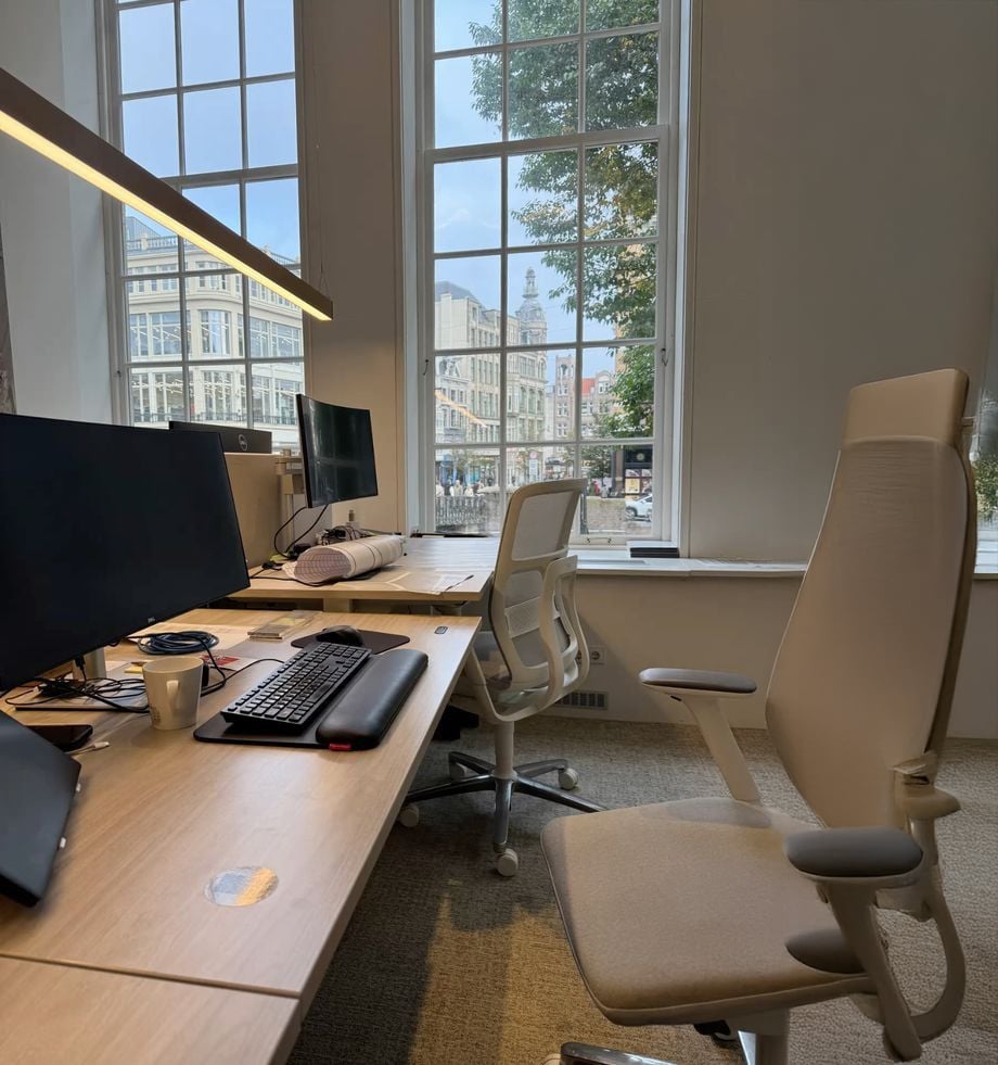 Modern office with two workstations, large windows with a city view, ergonomic chairs, computer monitors, a keyboard, and a coffee cup on the desk. Natural light fills the room.
