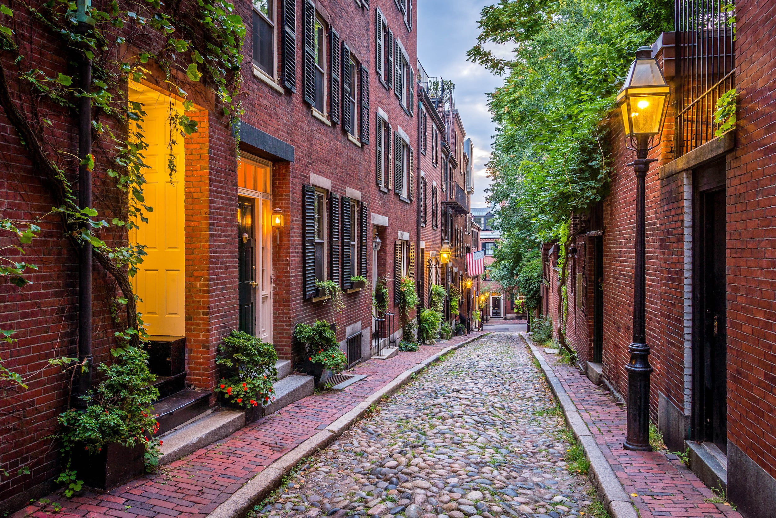 A narrow, cobblestone street lined with red brick buildings, black shutters, glowing lanterns, potted plants, and lush greenery, creating a charming, historic neighborhood scene at dusk.
