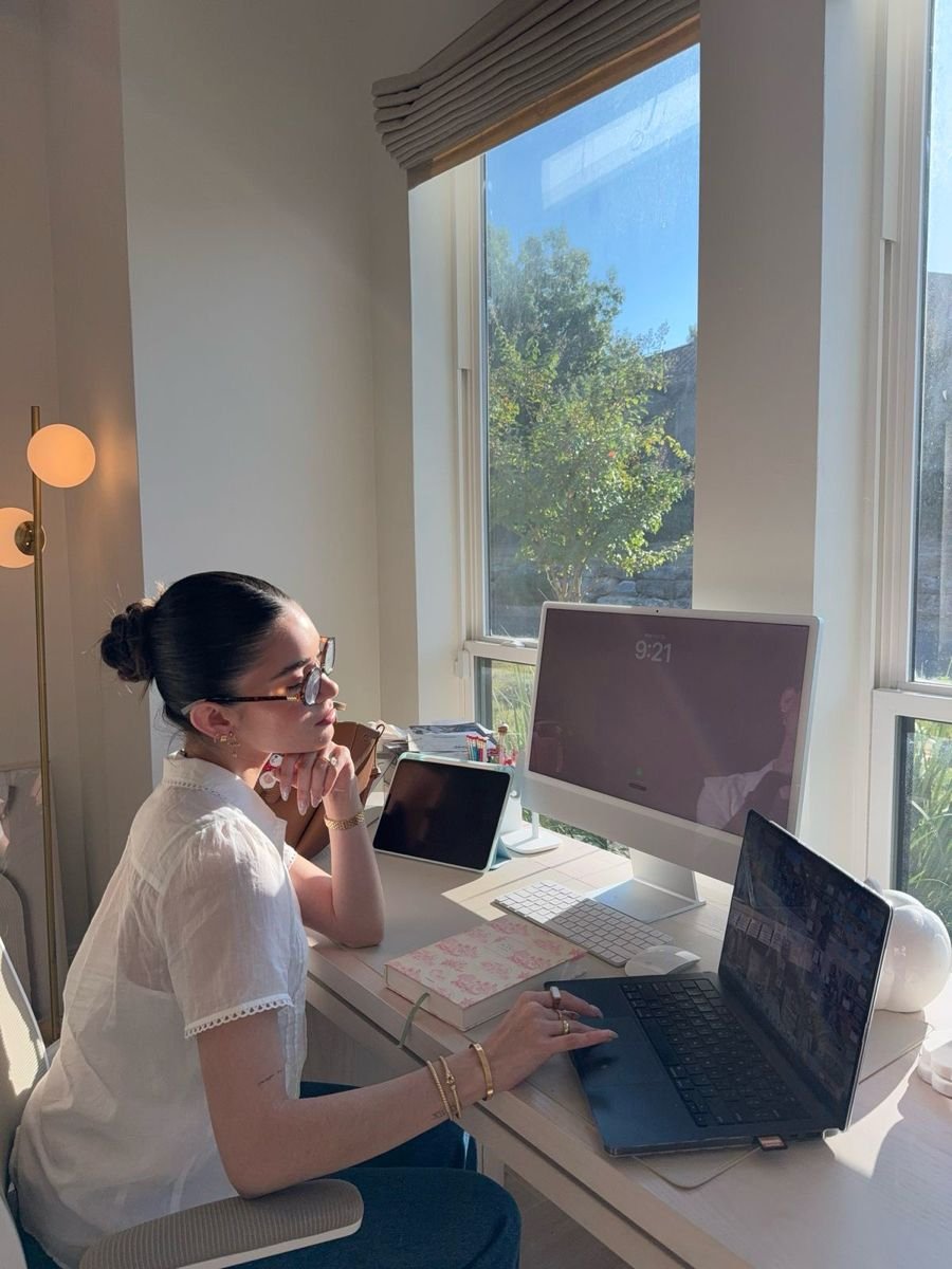 A woman sits at a desk by a sunny window, working on a laptop and desktop computer. She wears glasses and a white blouse, with notebooks and a tablet on the desk, and sunlight streaming into the room.