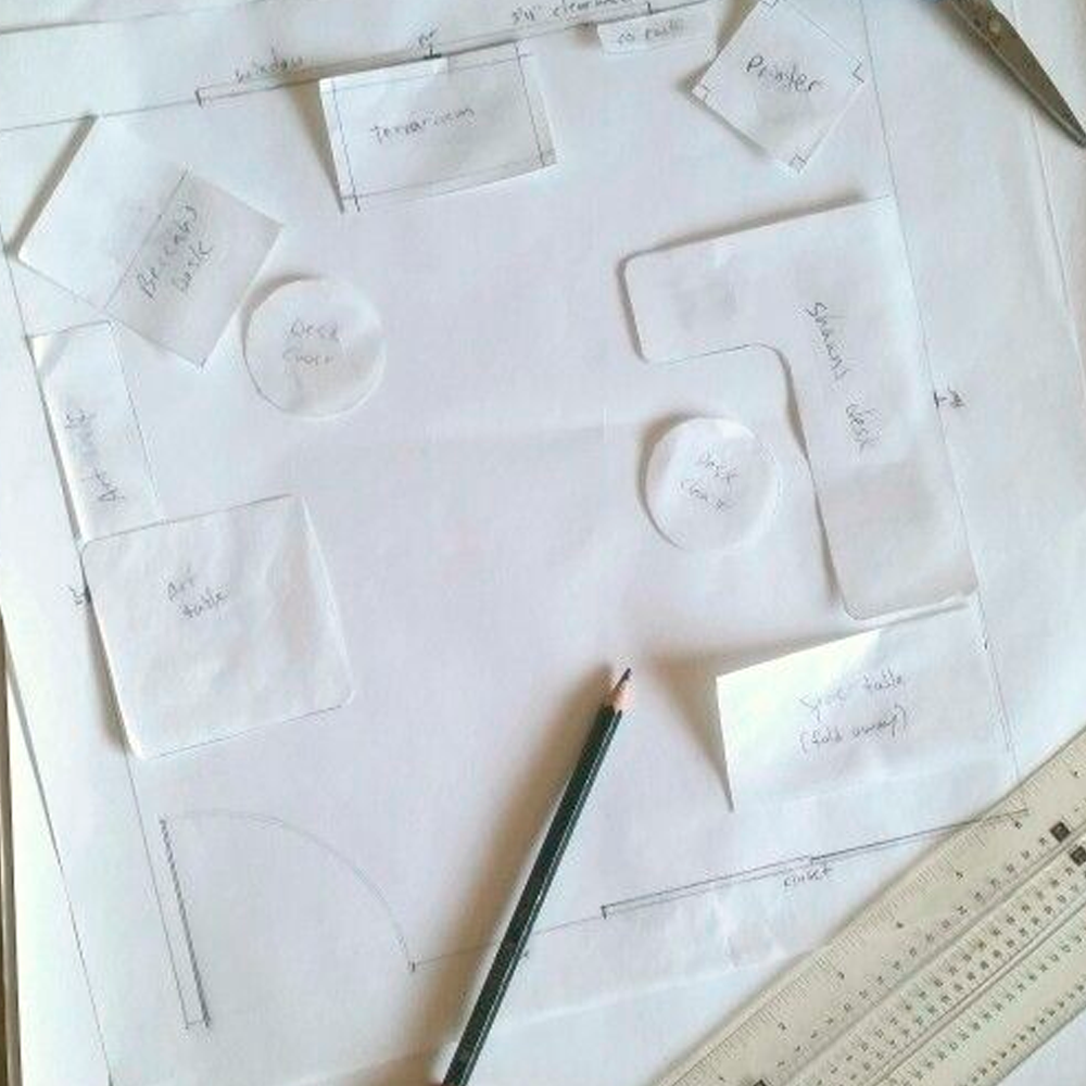 A workspace layout sketch on white paper with labeled paper cutouts for furniture, a pencil, a ruler, and handwritten notes. The design includes desks, an entrance, and various work zones.