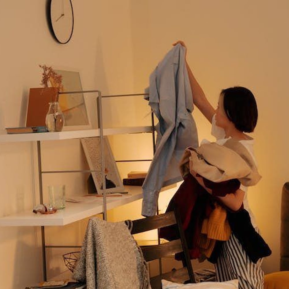 A person stands in a cozy, softly lit room holding several clothes, raising a blue shirt toward a shelf. A clock, books, and decorative items are arranged on the nearby shelves.